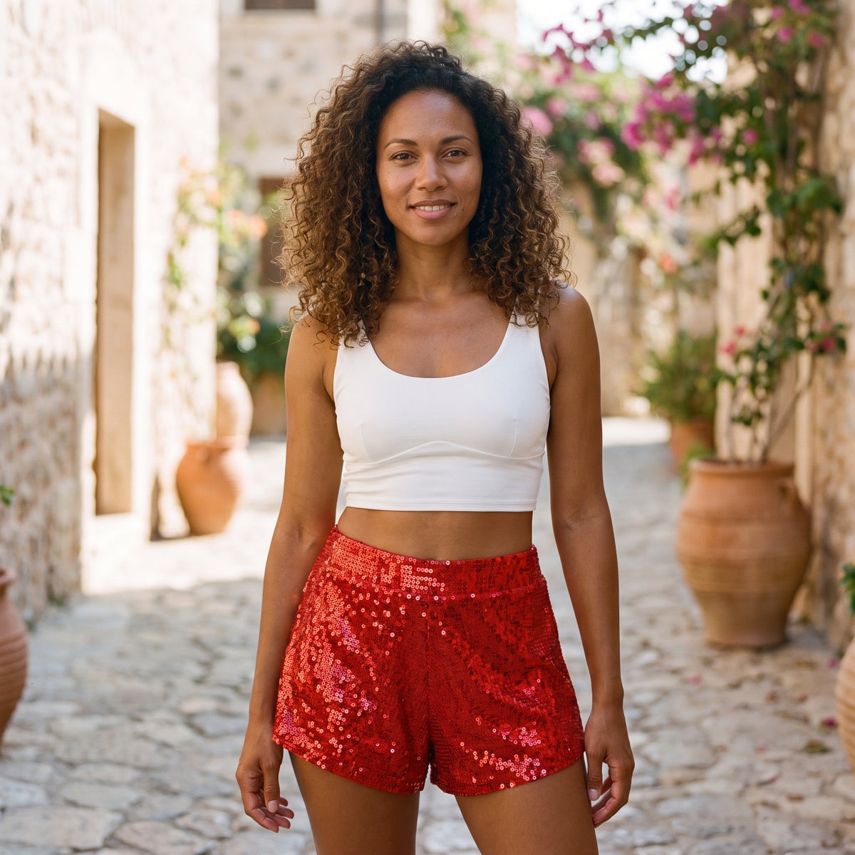 Modèle souriante portant un short à paillettes rouge taille haute, ruelle méditerranéenne pavée