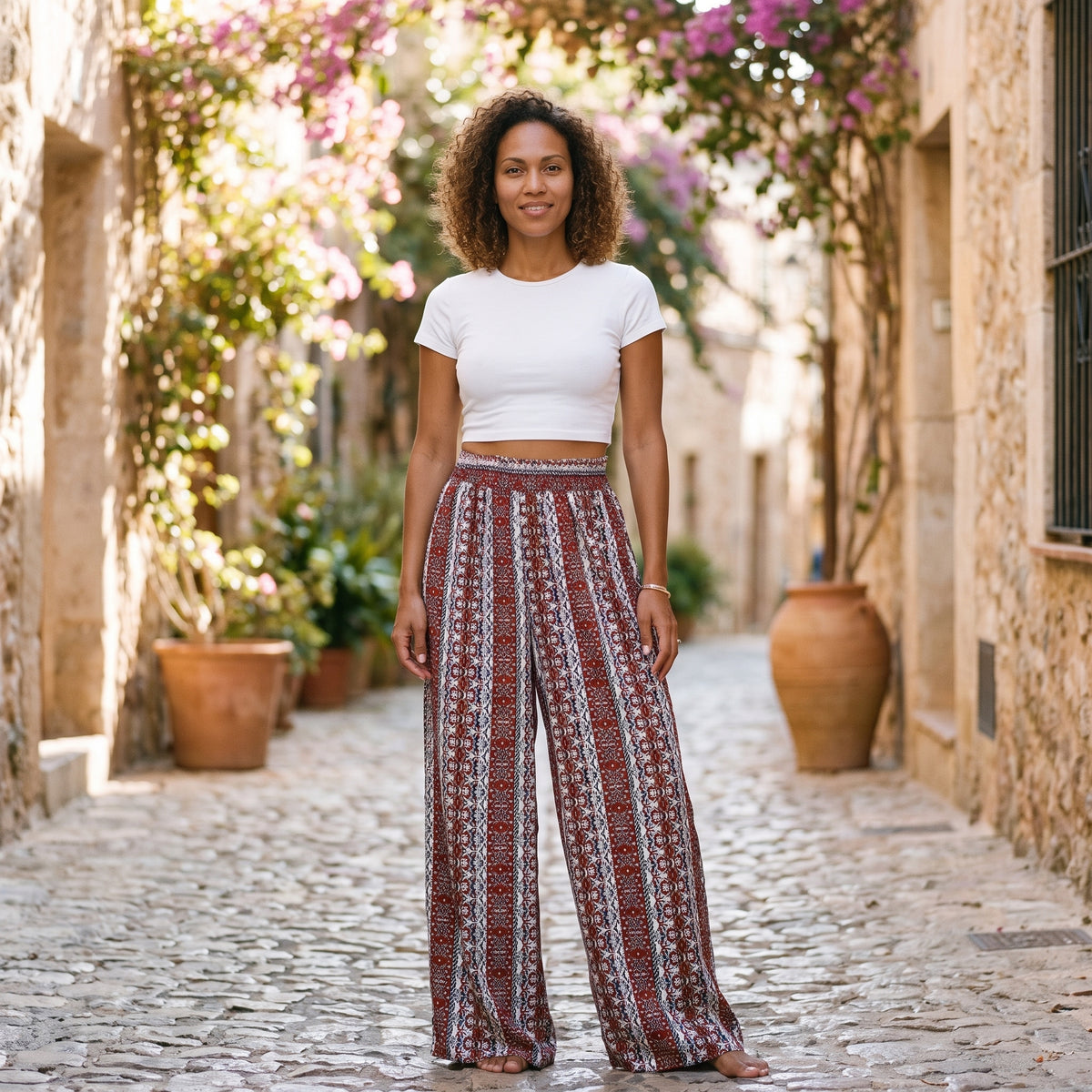 Modèle souriante portant le pantalon hippie large imprimé ethnique rouge et écru, taille haute, ruelle méditerranéenne pavée