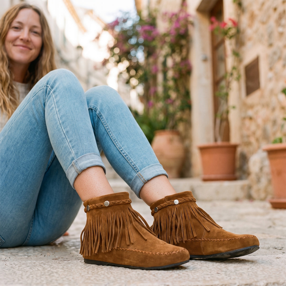 Modèle souriante portant les bottines mocassin à franges camel, assise dans une ruelle méditerranéenne