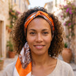 Modèle souriante portant le foulard cheveux hippie en satin orange à imprimé baroque, noué en bandeau dans les cheveux, ruelle méditerranéenne