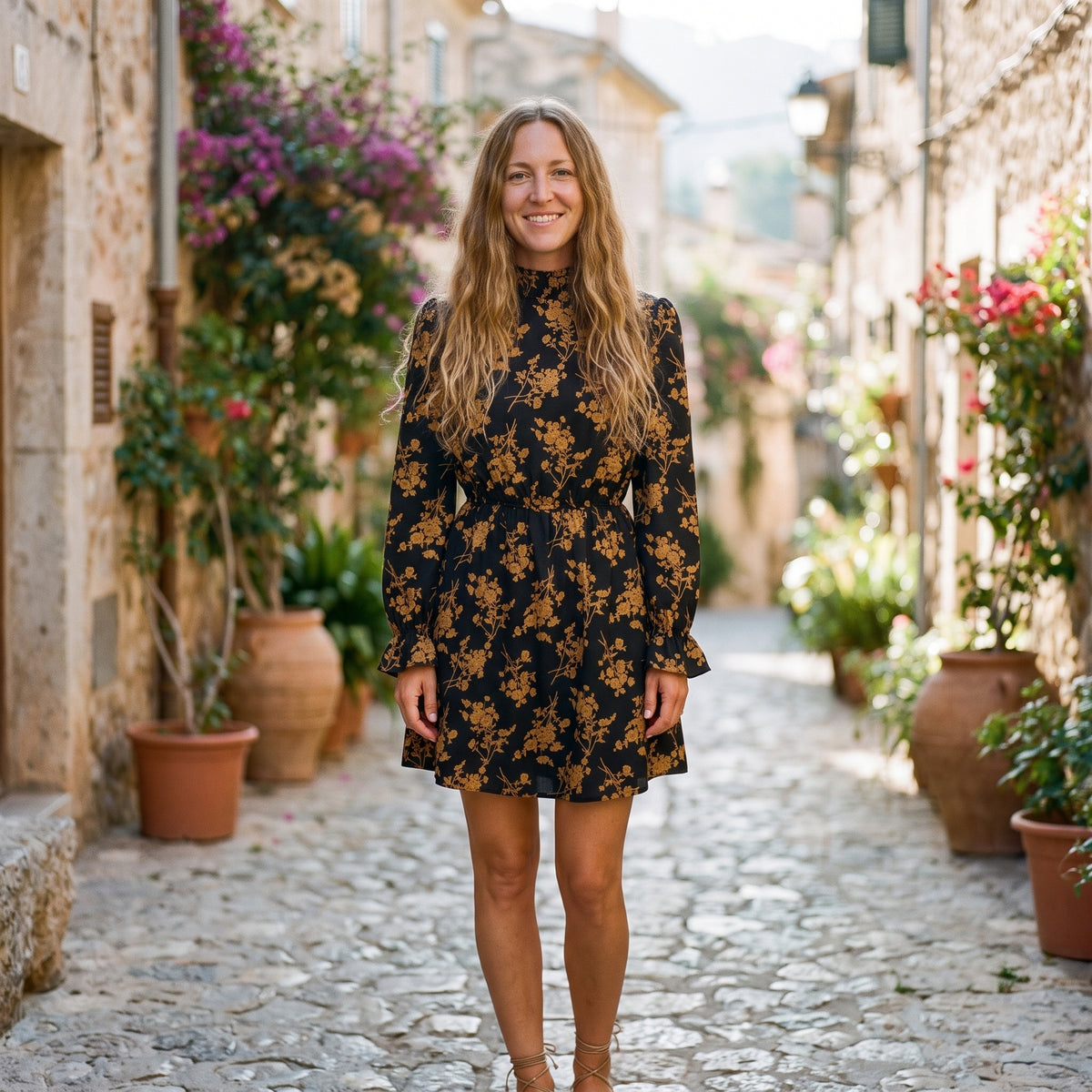 Modèle souriante portant la robe courte bohème noire à fleurs moutarde, ruelle méditerranéenne