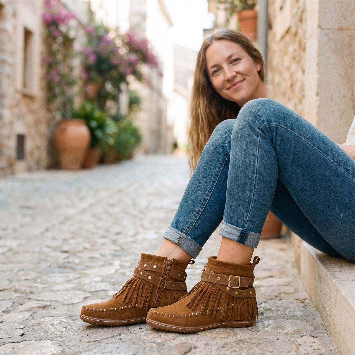 Modèle souriante portant les bottines mocassin à franges cloutées beige, ruelle méditerranéenne