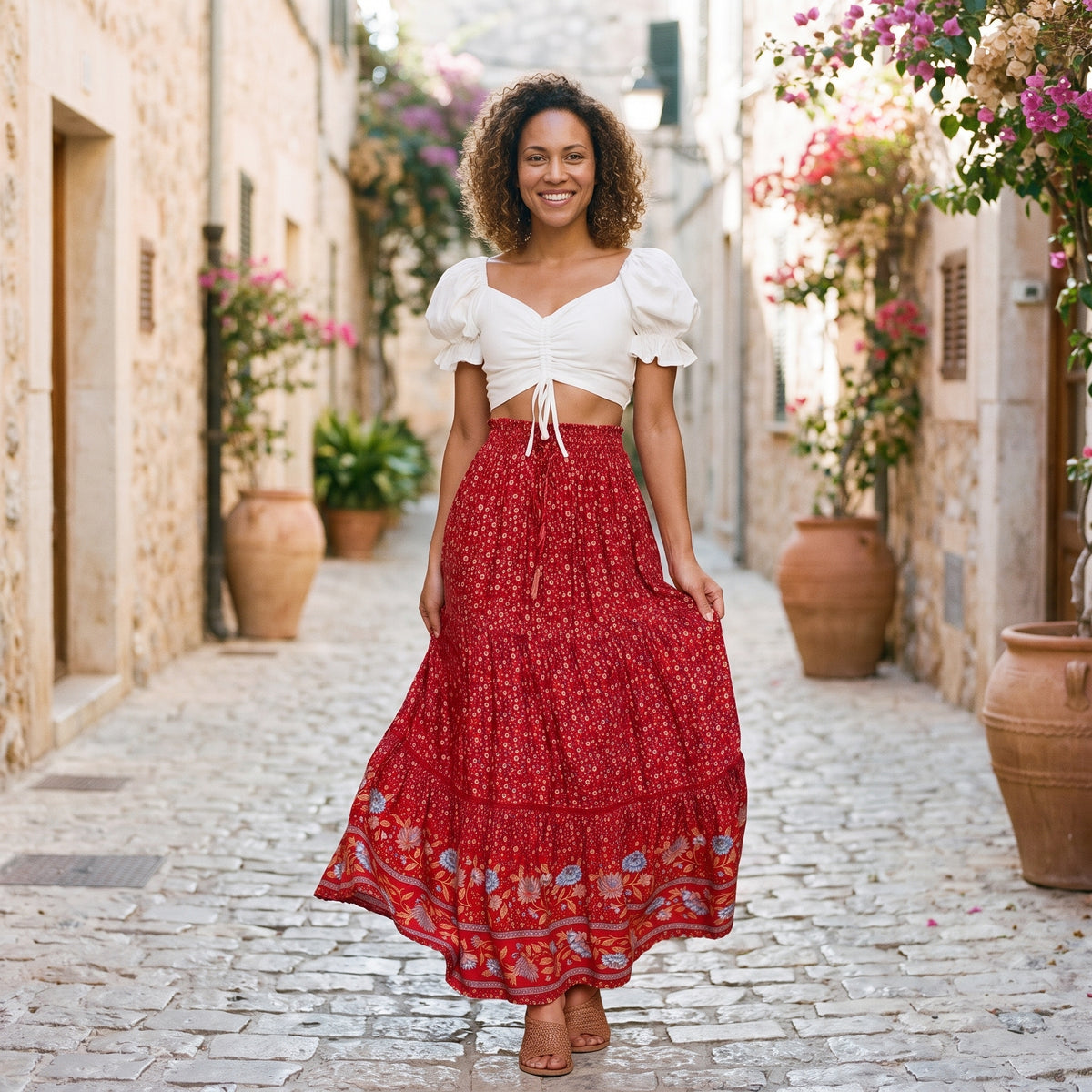 Modèle souriante portant la jupe hippie chic longue rouge à imprimé fleuri, debout dans une ruelle méditerranéenne ensoleillée